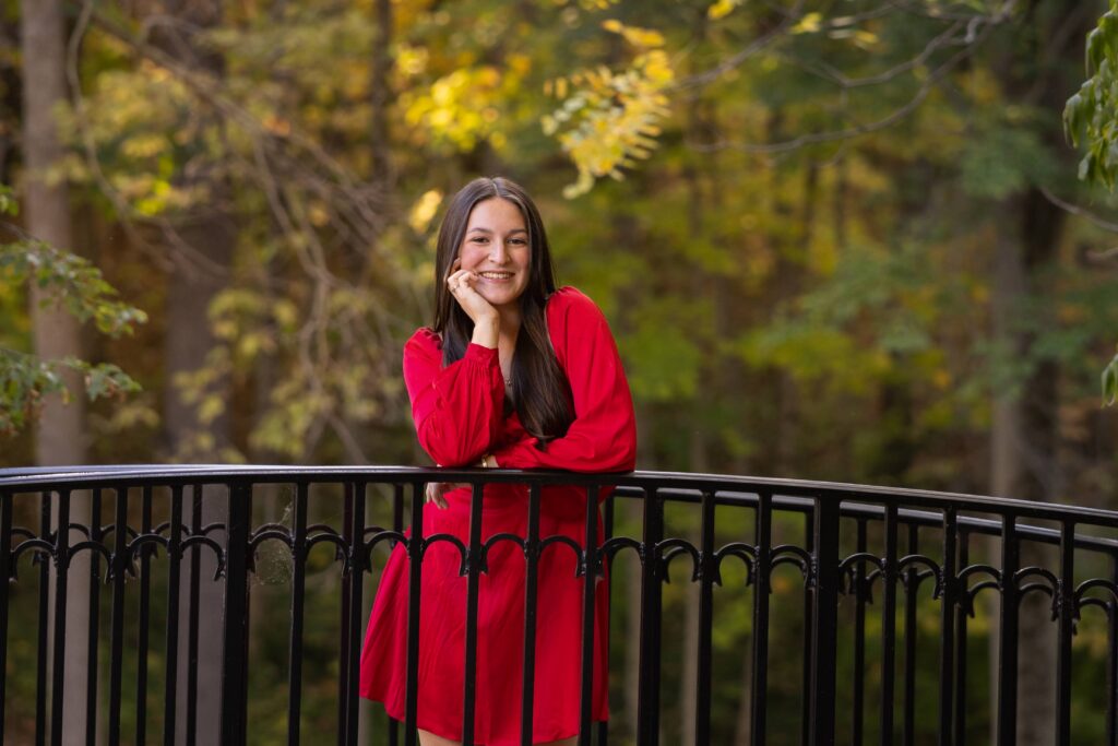 Senior portrait in a red dress leaning on an iron railing with fall foliage, Indianapolis senior photographer Dimensions Photography