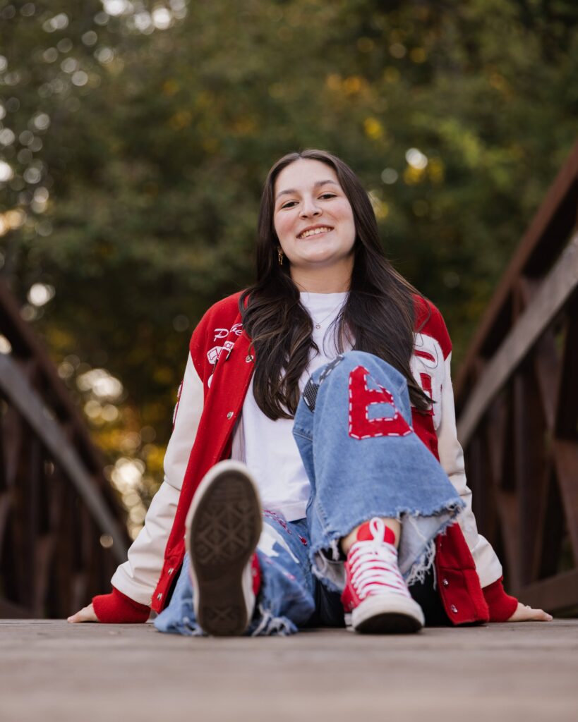 Senior wearing a red and white letterman jacket sitting on a wooden bridge, Indianapolis senior pictures by Dimensions Photography