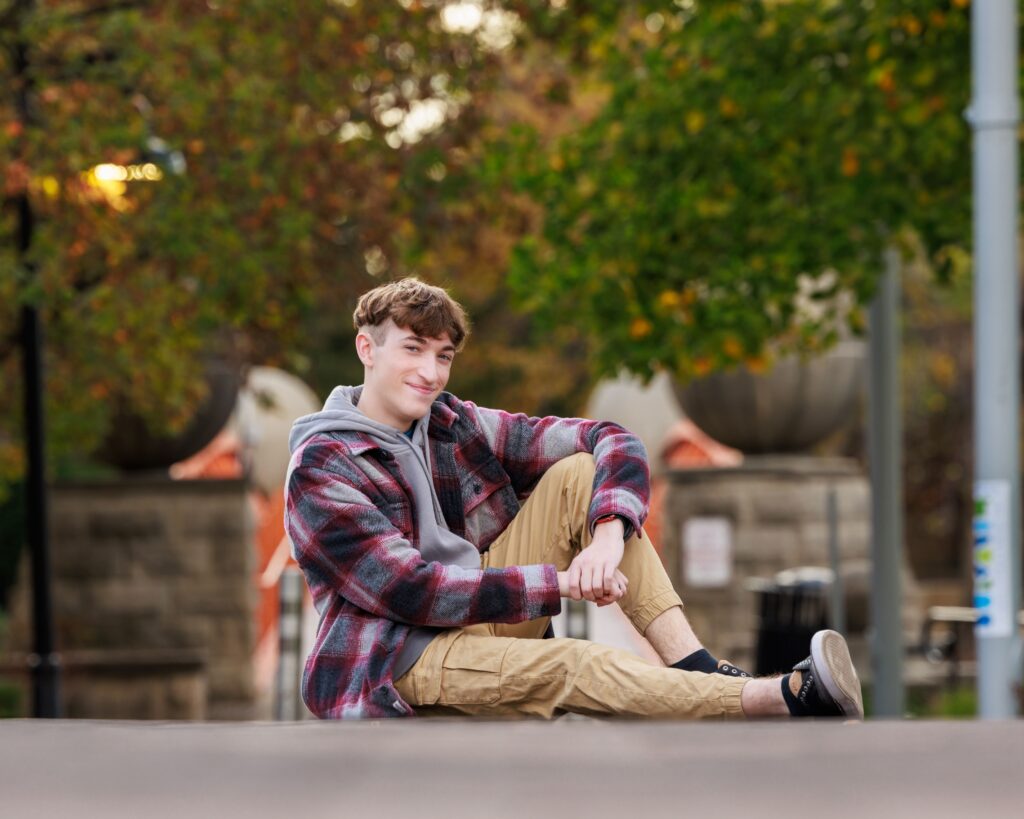 High school senior portrait of boy in plaid flannel jacket seated outdoors in park