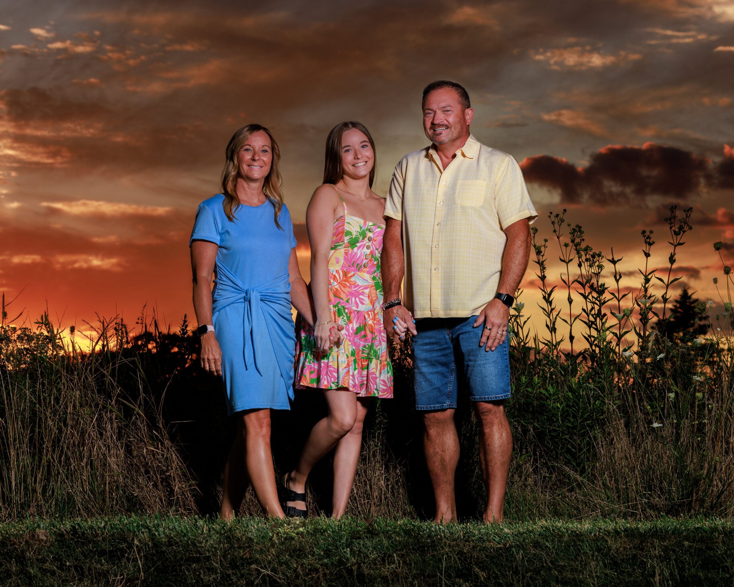 Family of three standing together in a field under a dramatic orange sunset sky