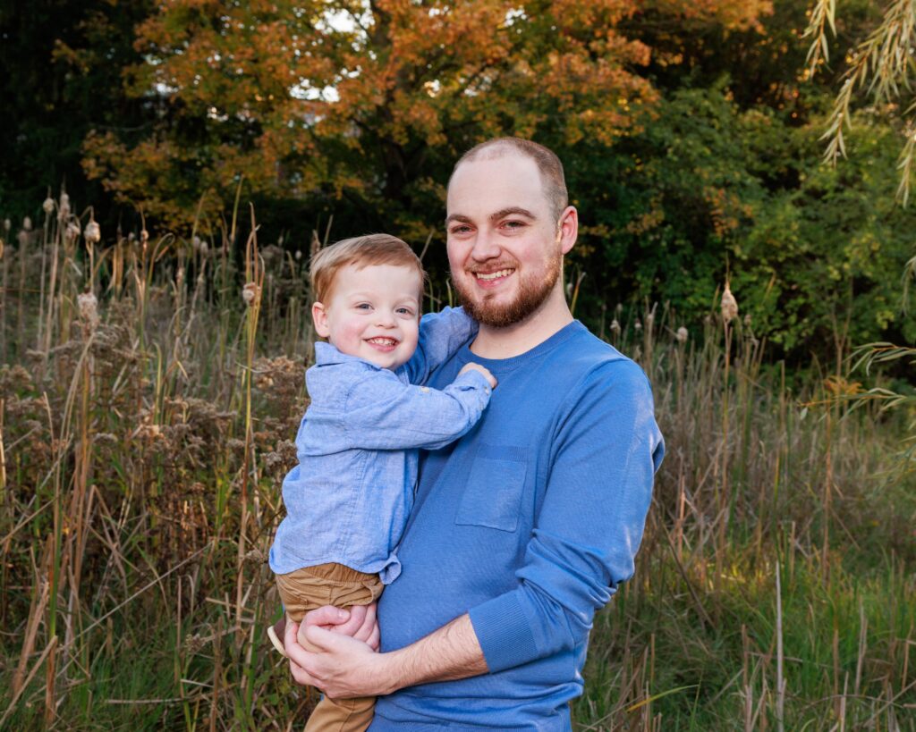 Father holding his toddler son in front of golden autumn trees during fall family photos in indianapolis