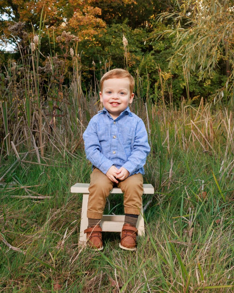 Toddler boy sitting on a small wooden bench surrounded by tall grass and fall foliage