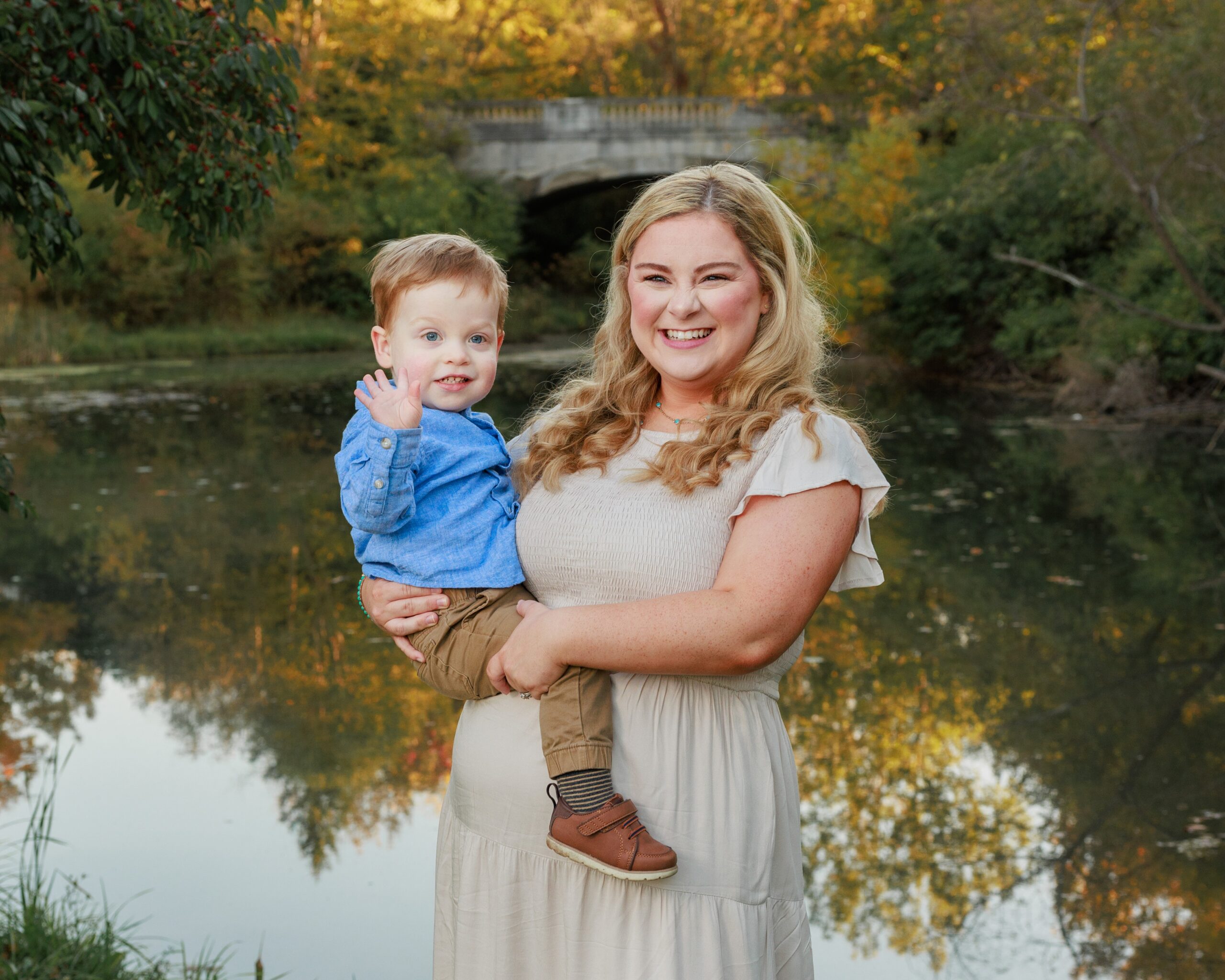 Mother holding her toddler son in front of a pond and stone bridge in autumn