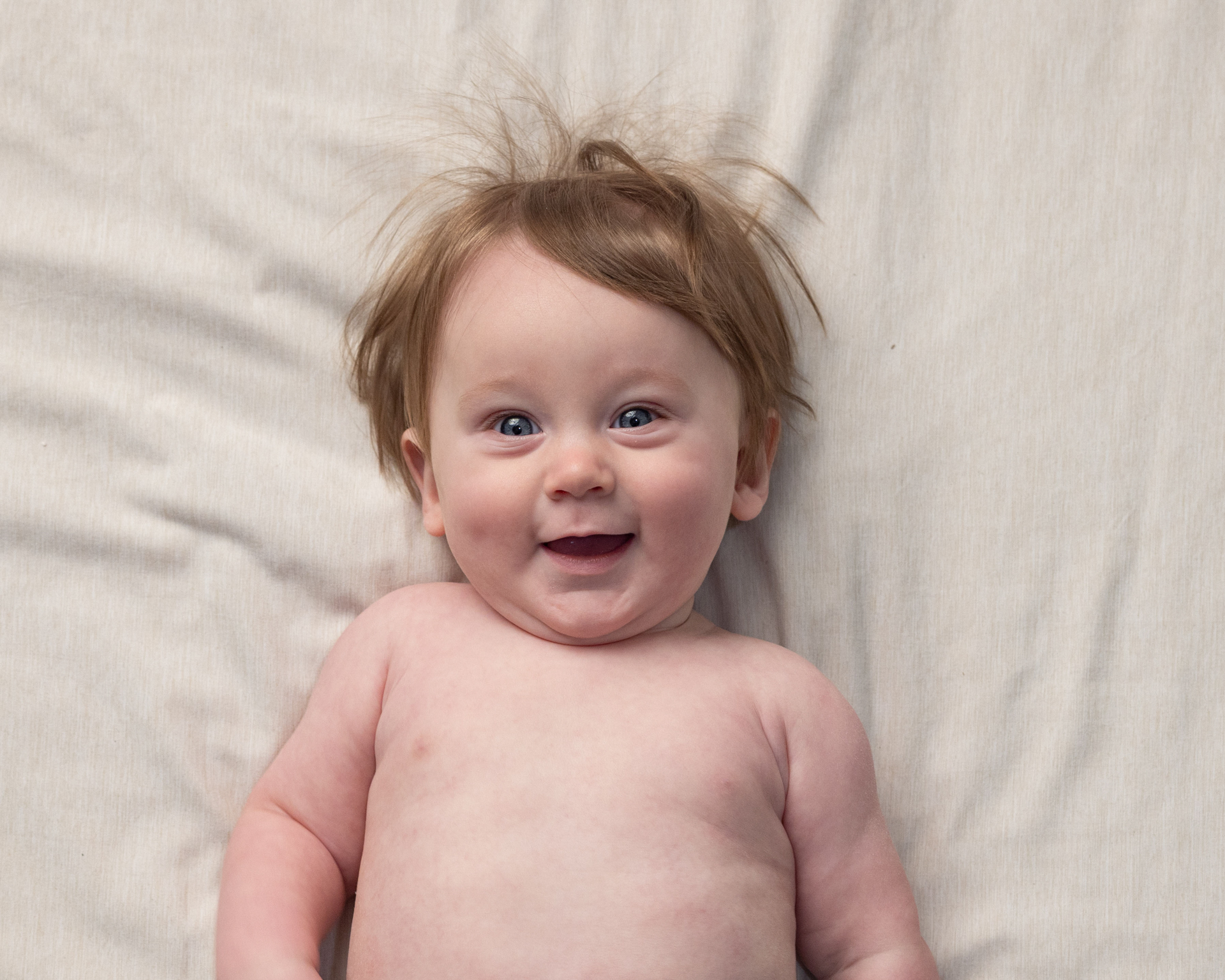 Baby giggling during a sitter session photography shoot, wispy hair backlit on cream fabric