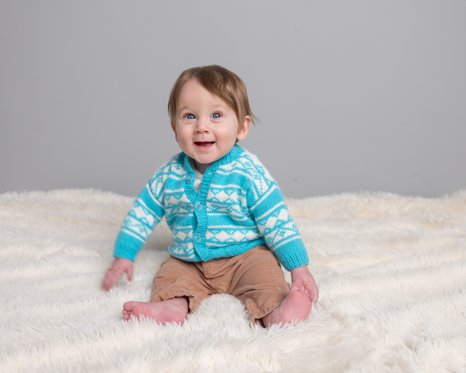Baby boy in a teal cardigan on a cream fur blanket, sitter session photography
