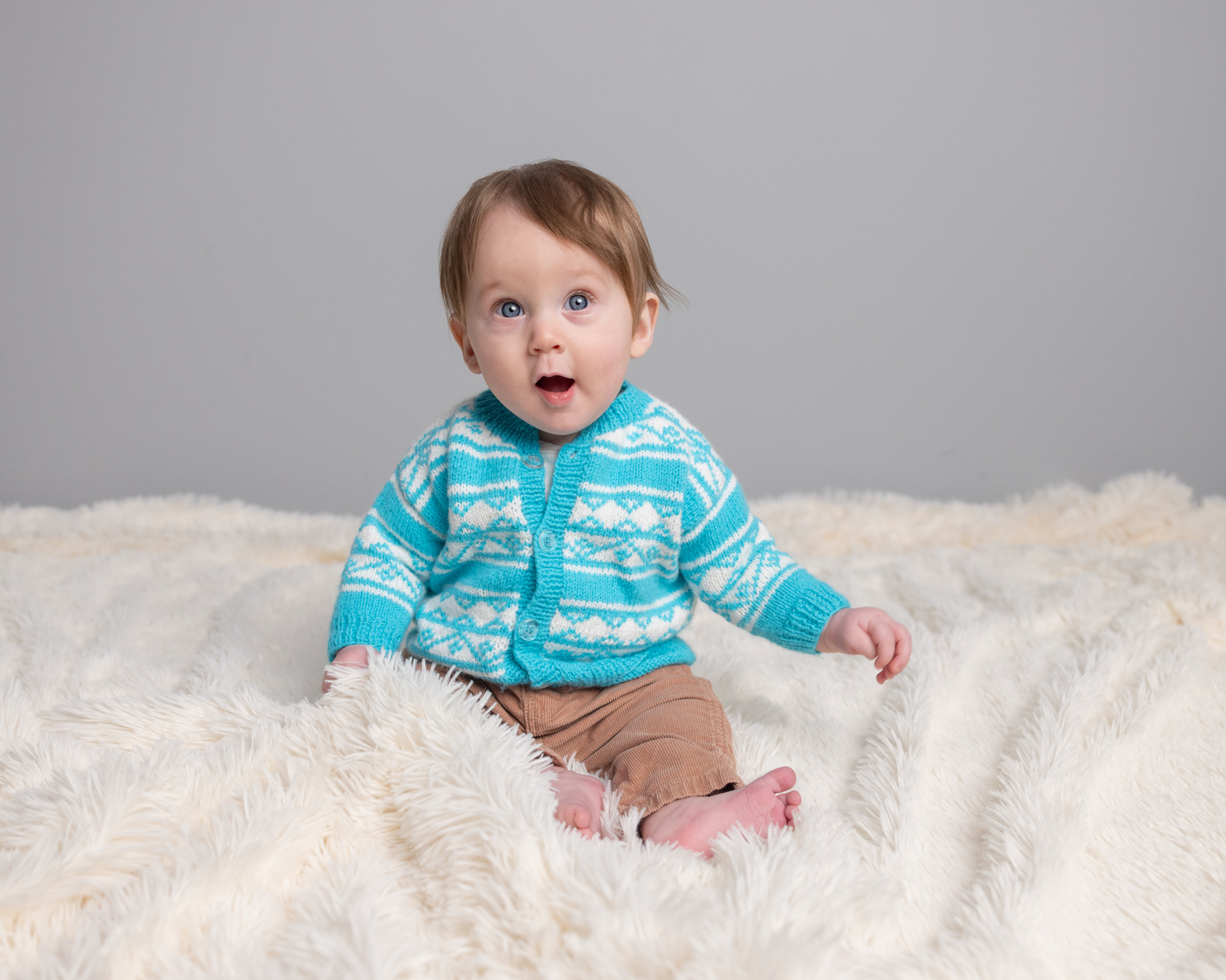 Baby boy in teal cardigan with a surprised, open-mouth expression, sitter session photography