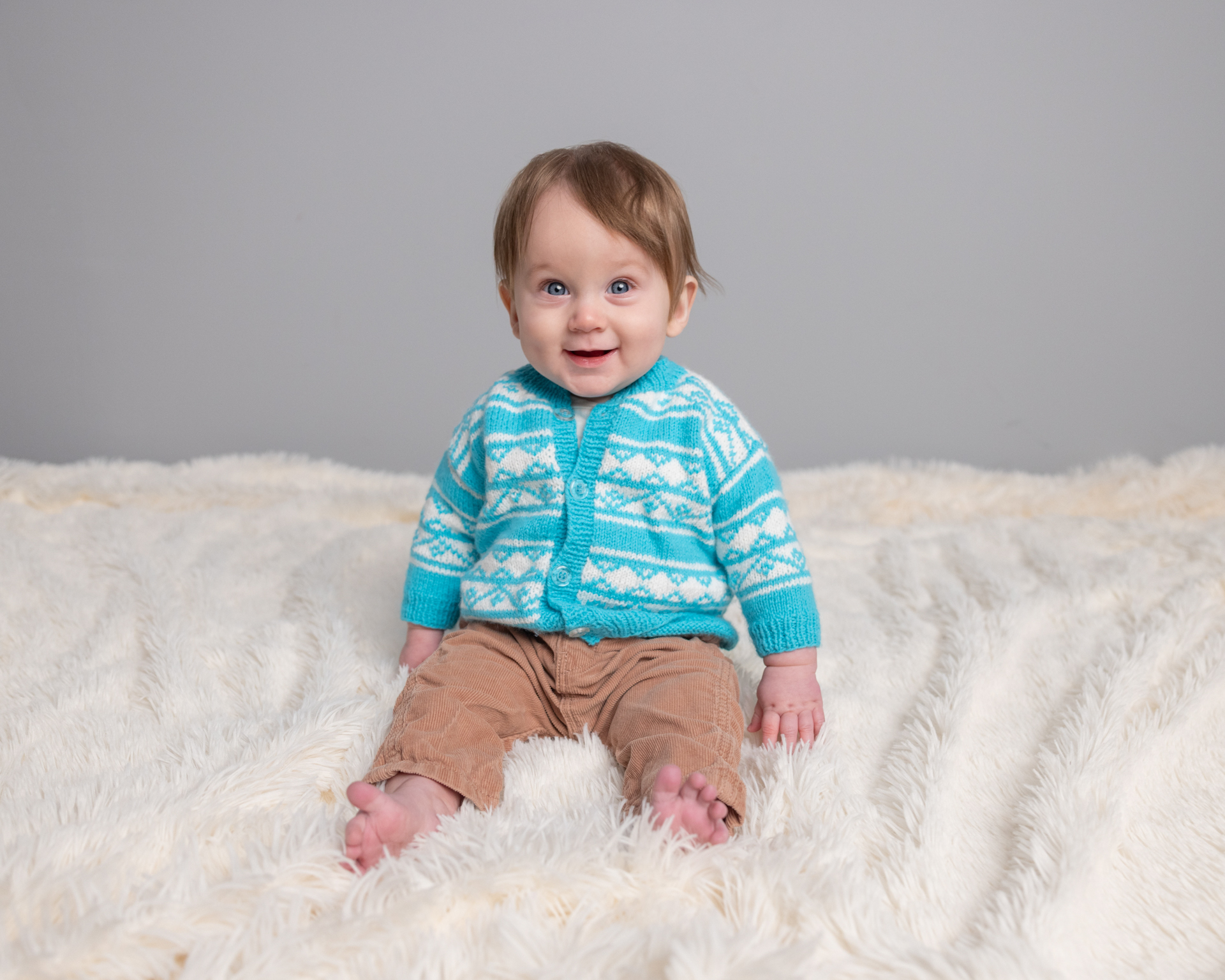 Baby boy in a teal cardigan on a cream fur blanket, sitter session photography