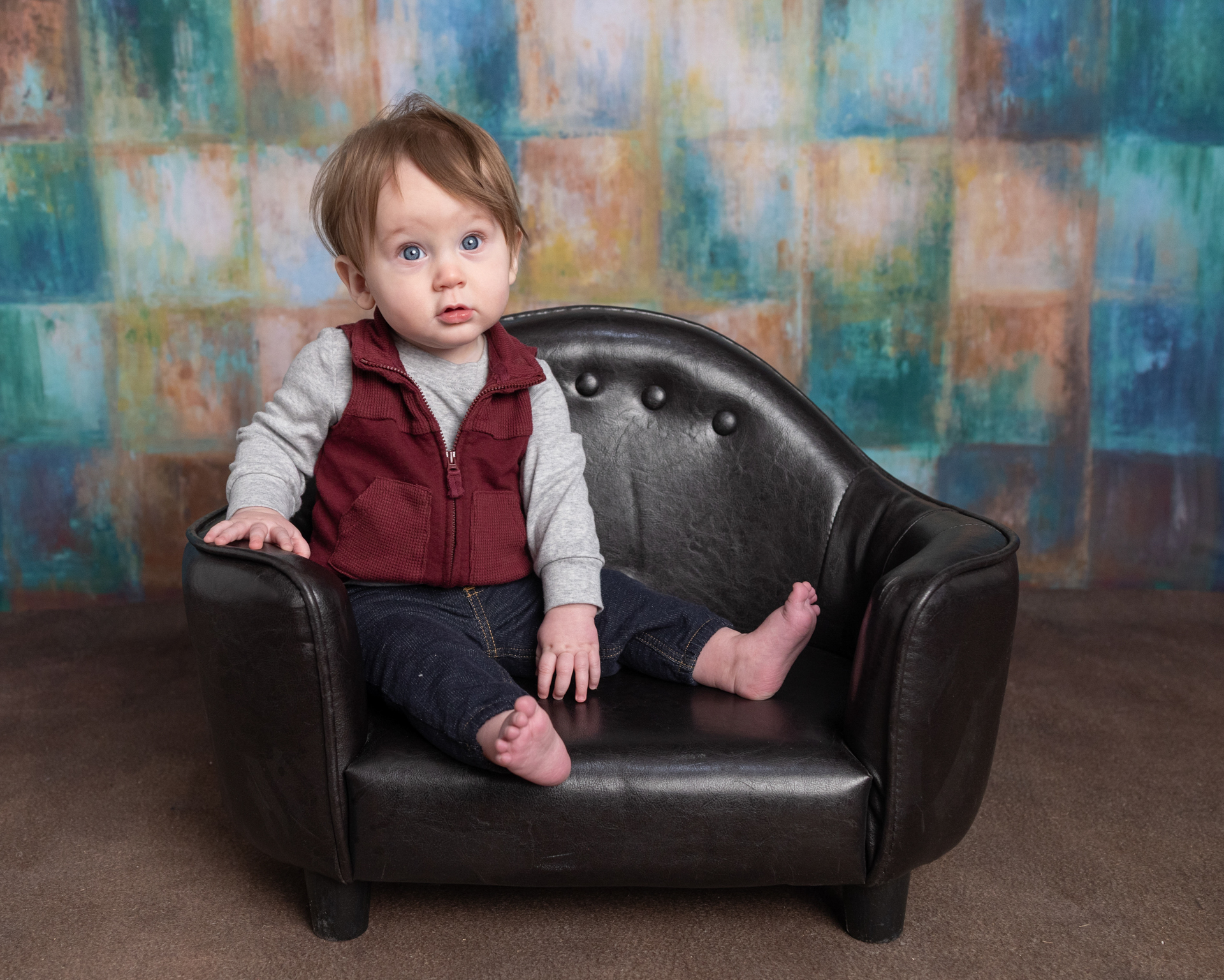 Baby boy in a maroon vest sitting in a leather mini chair