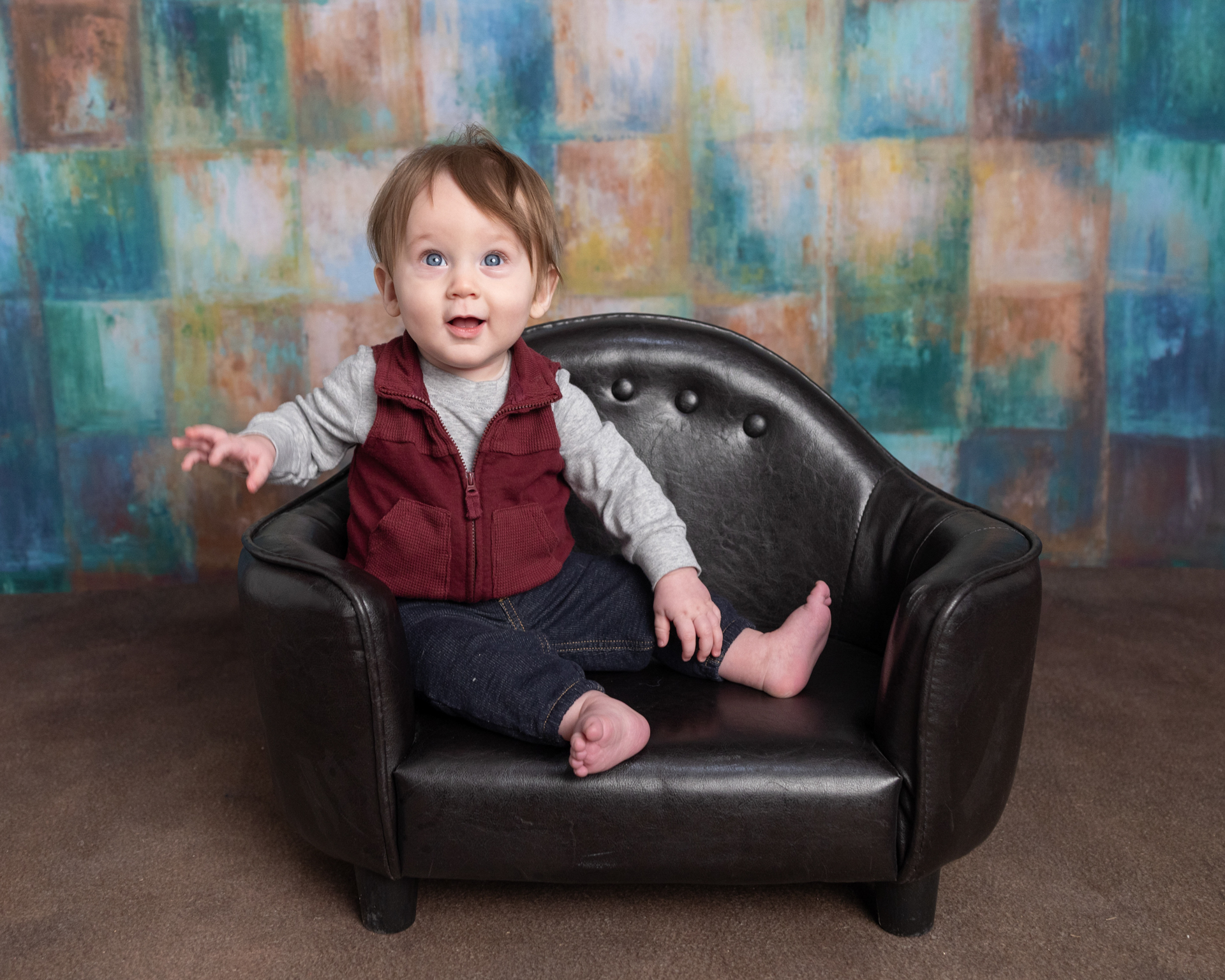 Baby boy reaching out from a leather mini chair against a colorful painted backdrop