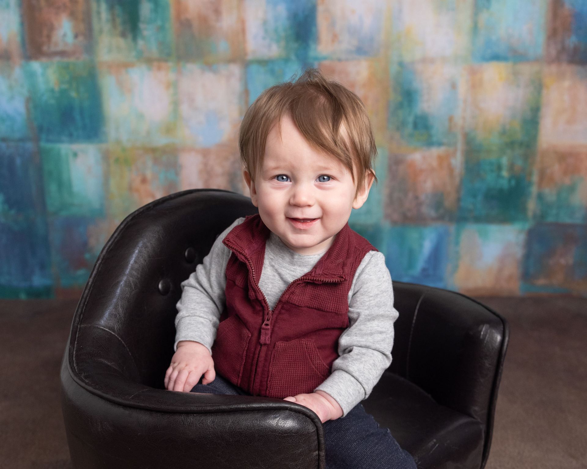 Baby boy smiling in a leather mini chair during his sitter session photography portrait